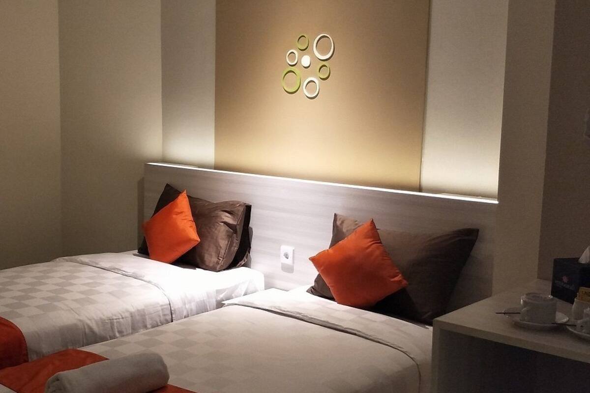 Quarto de hotel com duas camas, cada uma com colcha branca e laranja, e um candeeiro fixado na parede sobre as camas.