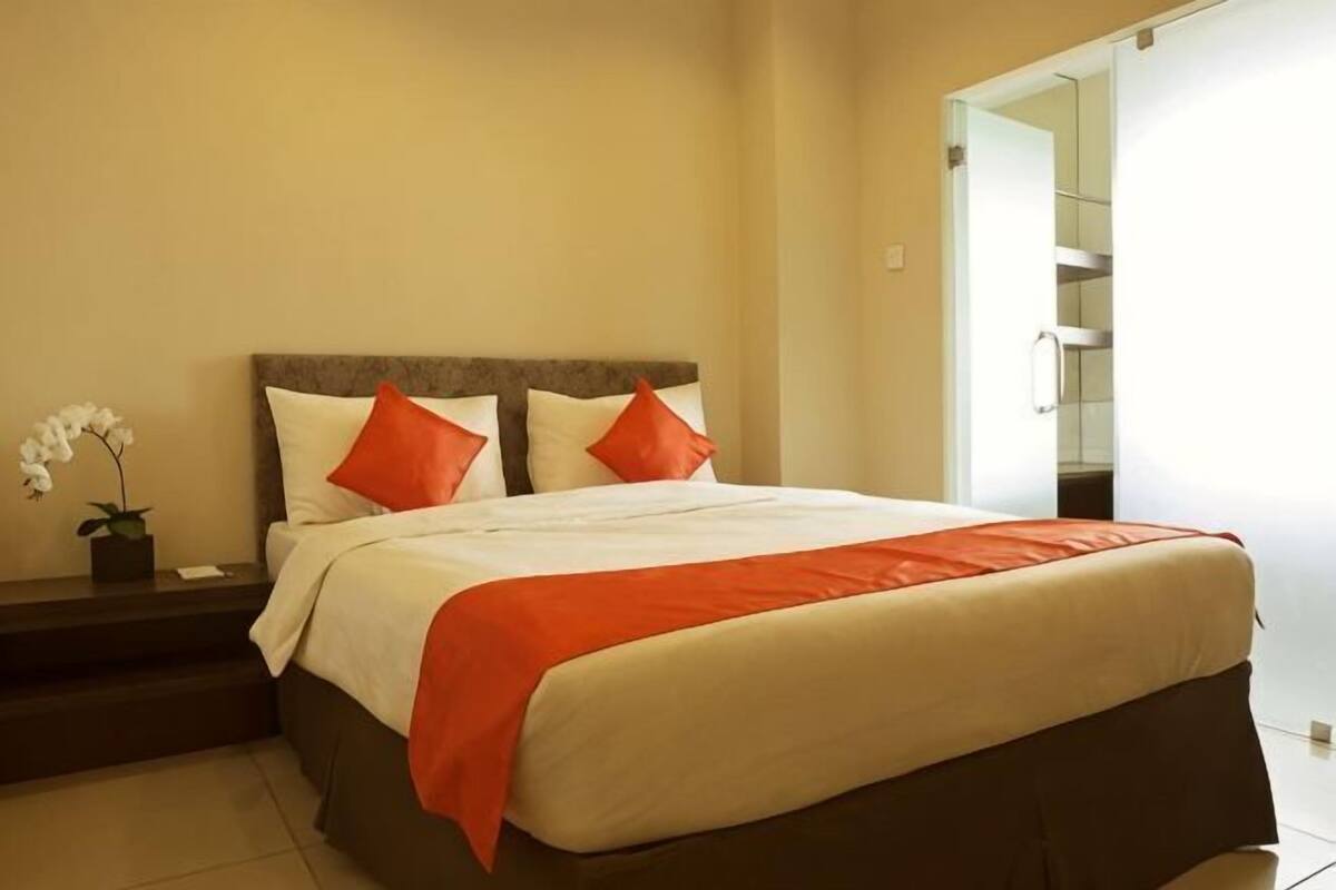 Quarto de hotel com cama, almofadas, mesa de cabeceira e planta em vaso.