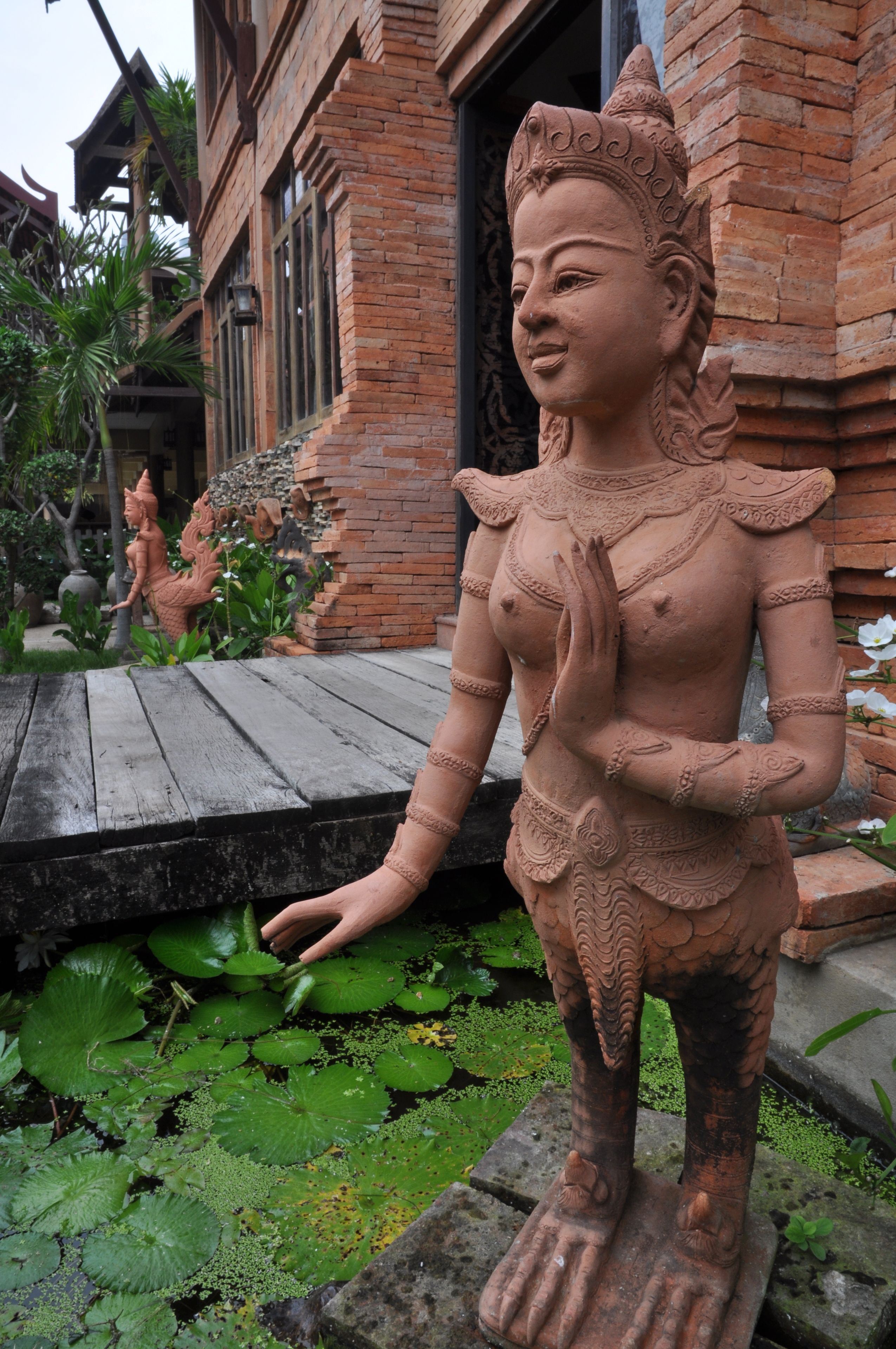 Una estatua de terracota de una deidad en un jardín, con un edificio de ladrillo al fondo.