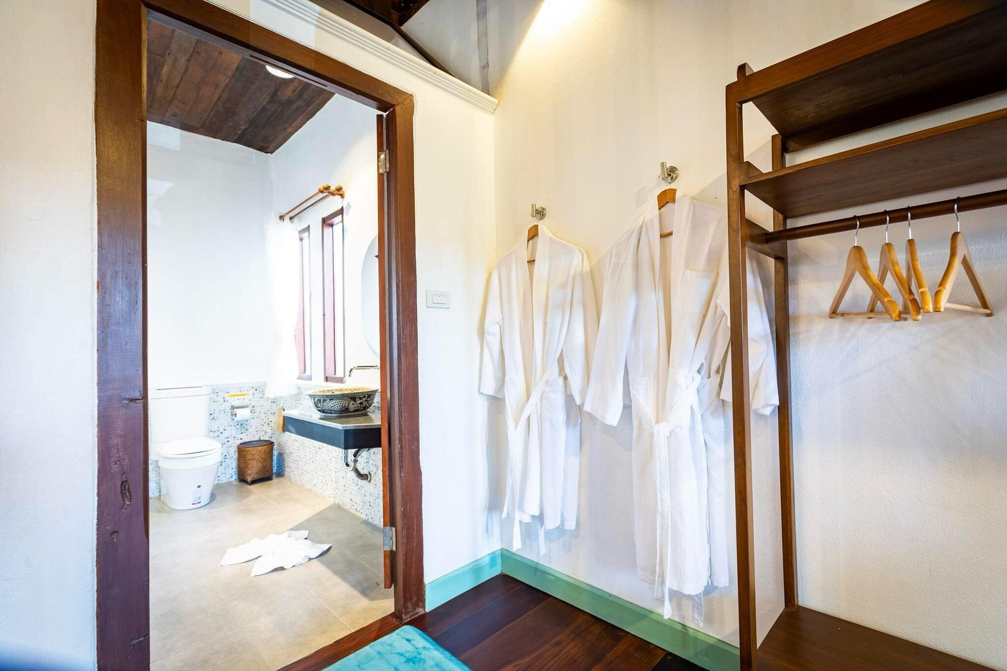 A bathroom with a wooden door, a sink, and a mirror. There are white robes hanging on a rack.