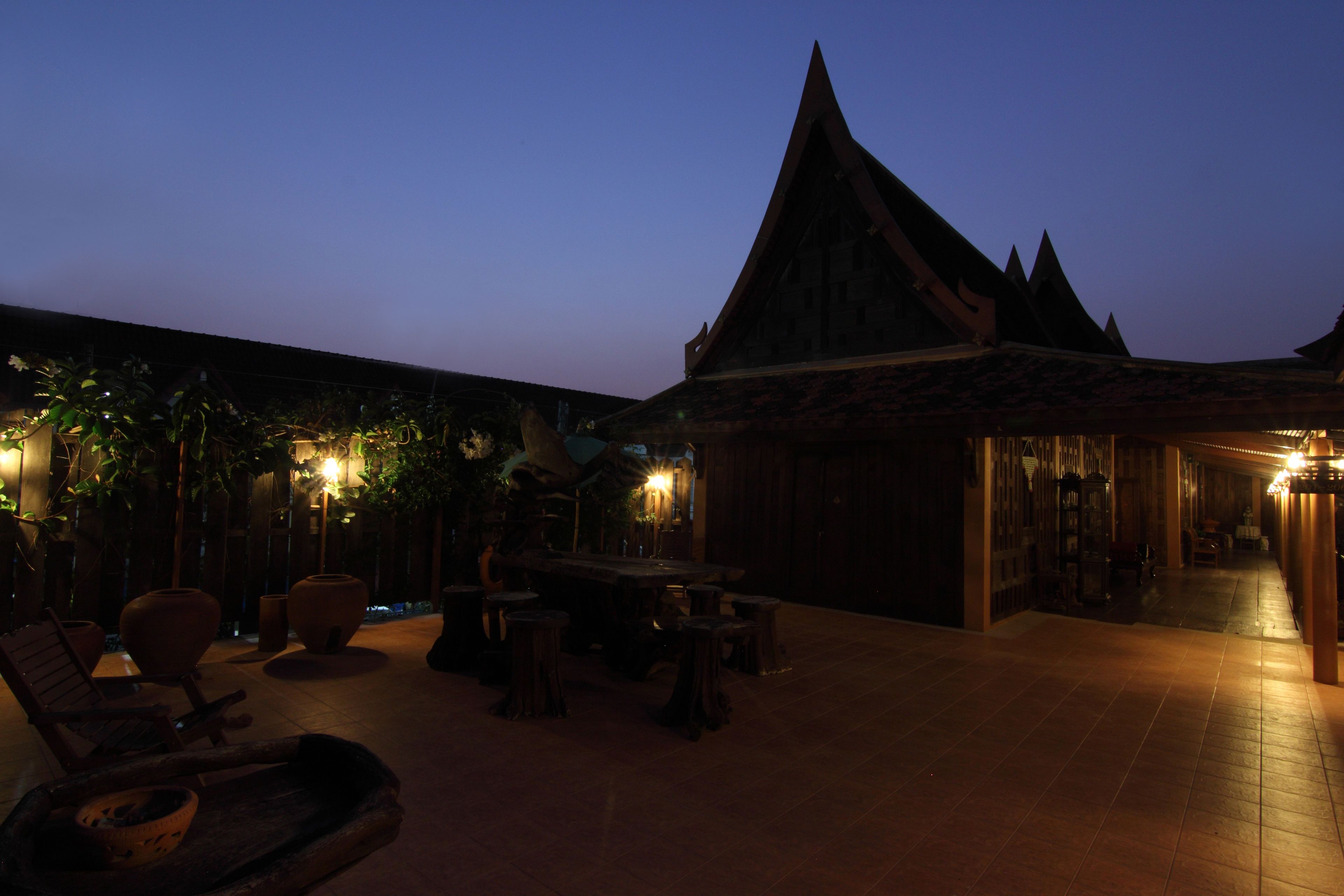 An outdoor seating area with a wooden table and chairs, surrounded by potted plants and a building with a pointed roof.