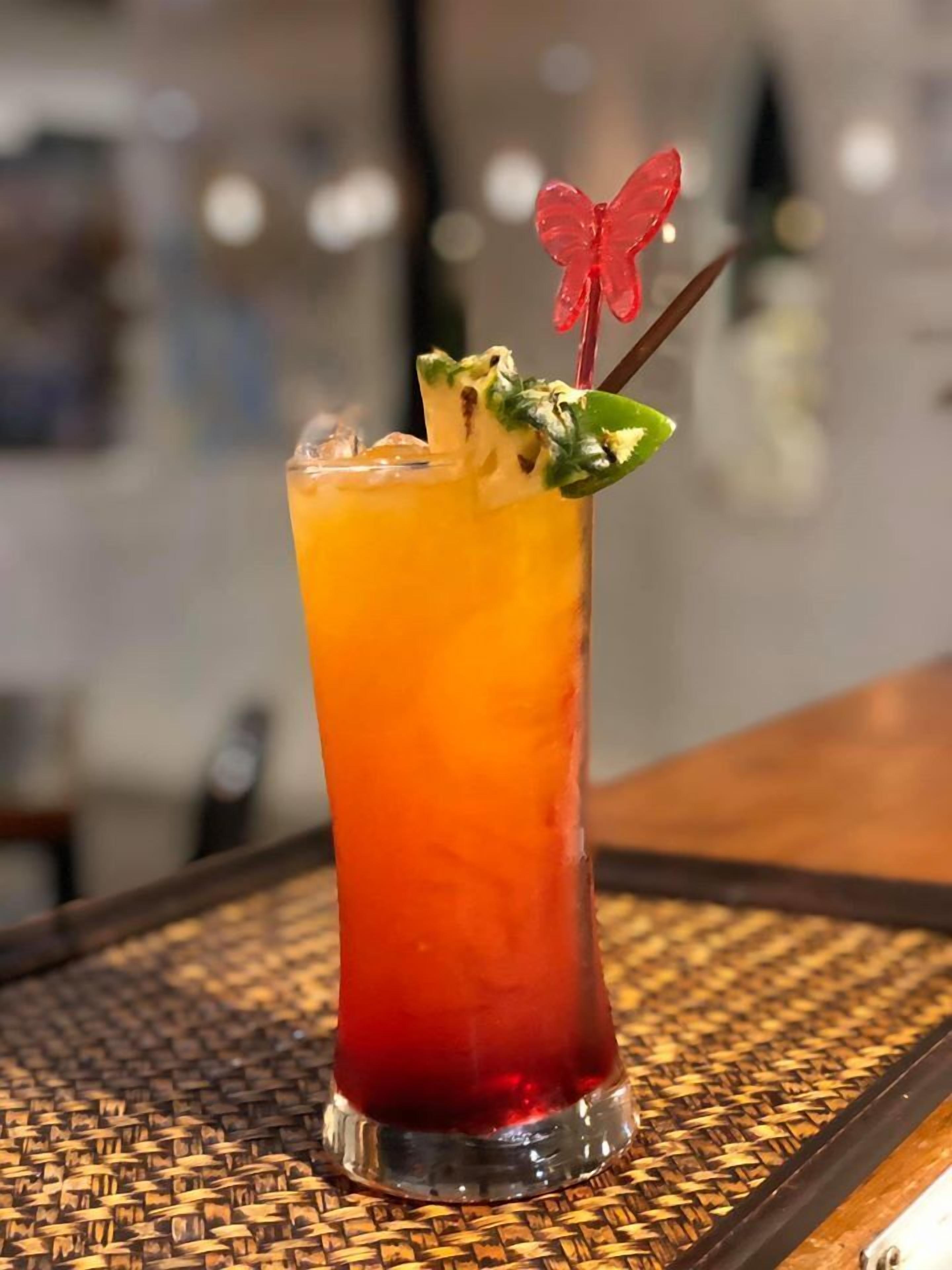 A tall glass with a gradient drink, garnished with a red butterfly and mint leaves, on a woven mat.