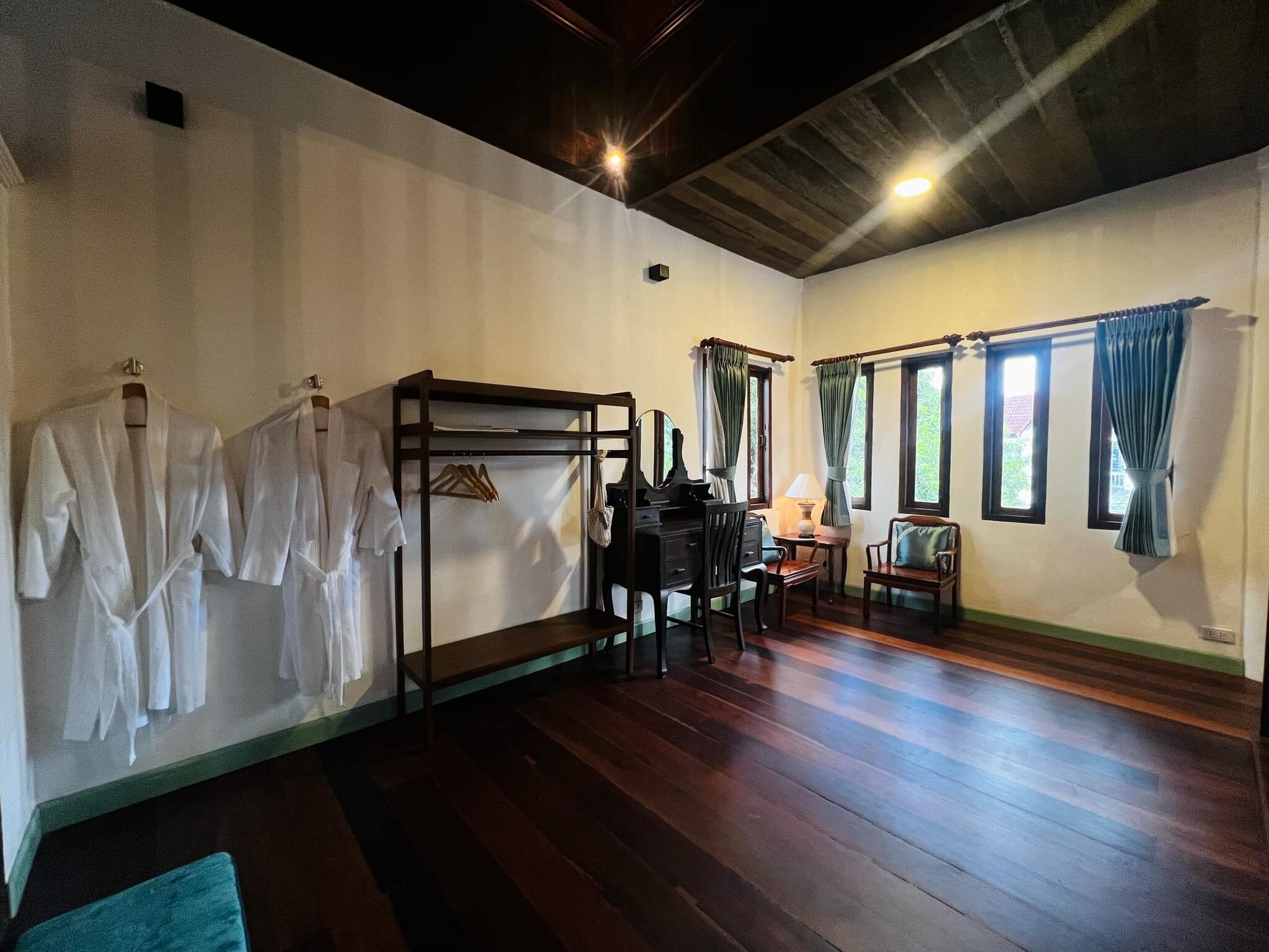 A room with wooden flooring, a black desk with a chair, a small table with a lamp, and a wardrobe with clothes hanging.