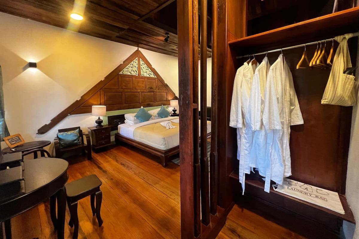 A bedroom with a wooden ceiling, a bed with white linens, a nightstand, a desk with a chair, and a wardrobe with hanging clothes.