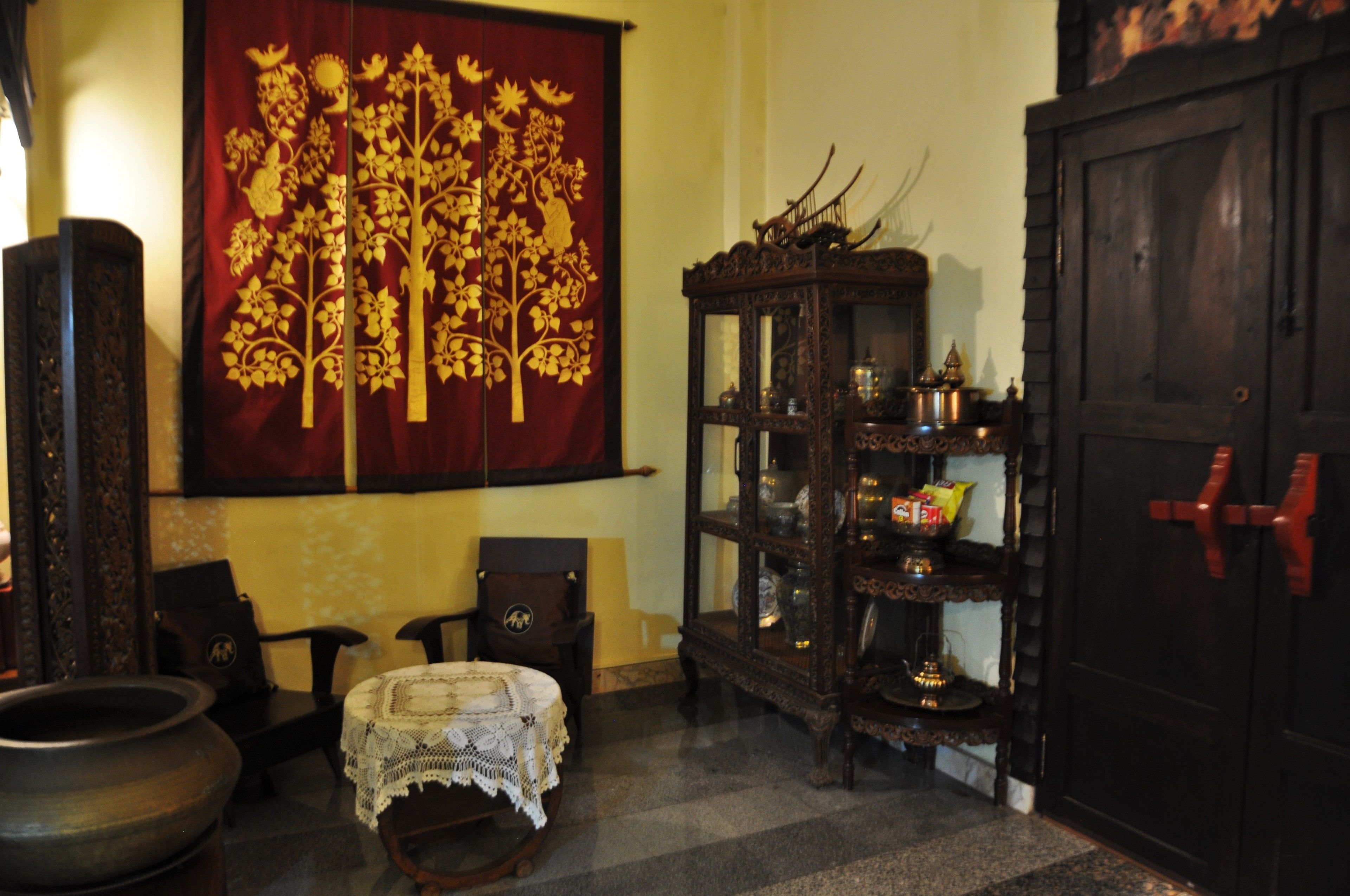 A traditional room with a large ornate tapestry, a wooden cabinet with glass doors, and a dark wooden door with a red handle.