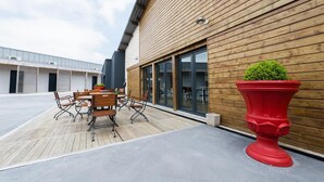 An outdoor patio with a wooden table and chairs, a red planter with a small tree, and a building with large windows and a wooden facade. - Le Relais Du Boisniard (Chanverrie)