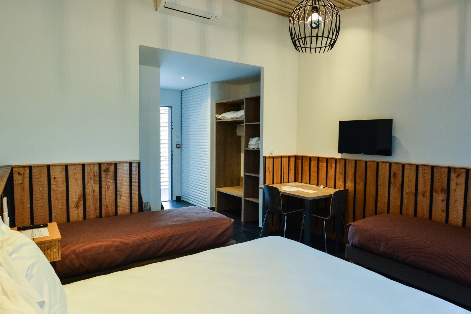 A hotel room with two single beds, a small table with chairs, a TV mounted on the wall, and a wardrobe with shelves.