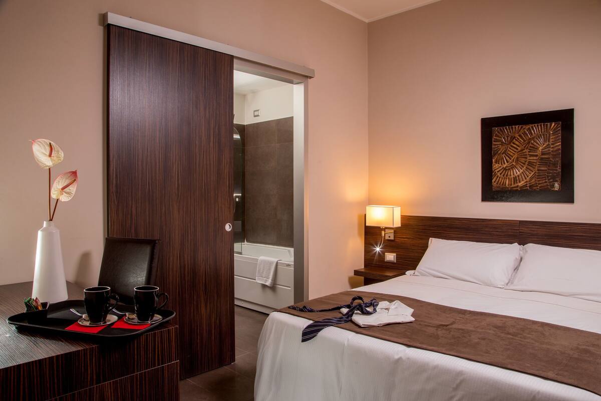 Habitació d'hotel amb un llit, un escriptori amb dues tasses i una safata, un bany amb banyera i una obra d'art penjada a la paret.