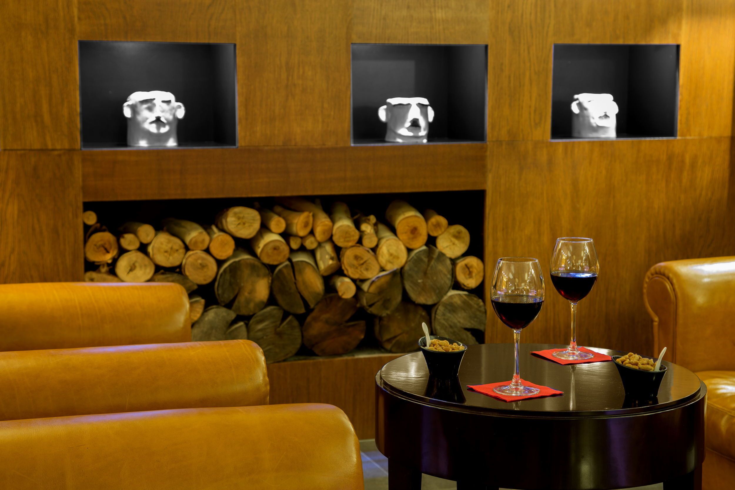 Un coin salon accueillant avec des fauteuils en cuir, une table ronde sur laquelle reposent deux verres à vin et un bol de céréales, et un mur décoré de rondins de bois et de trois objets encadrés.