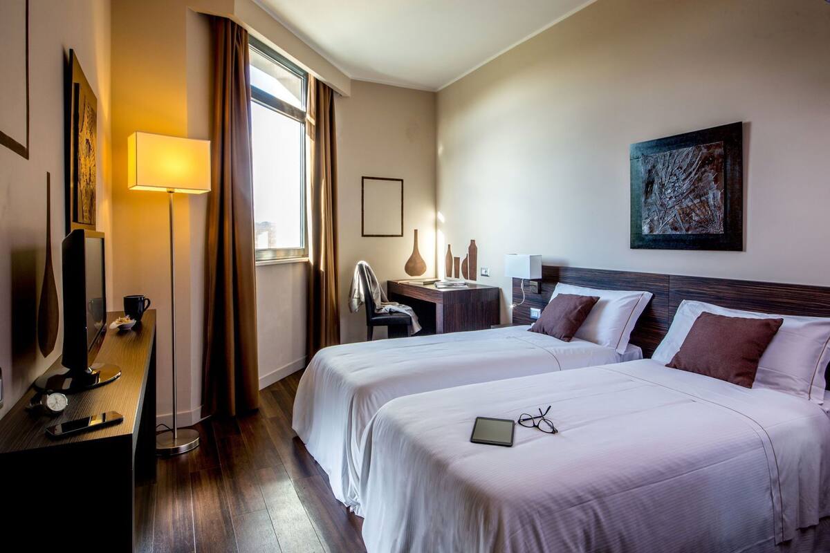 Habitació d'hotel amb dos llits, un escriptori amb un ordinador, una làmpada i un quadre a la paret.