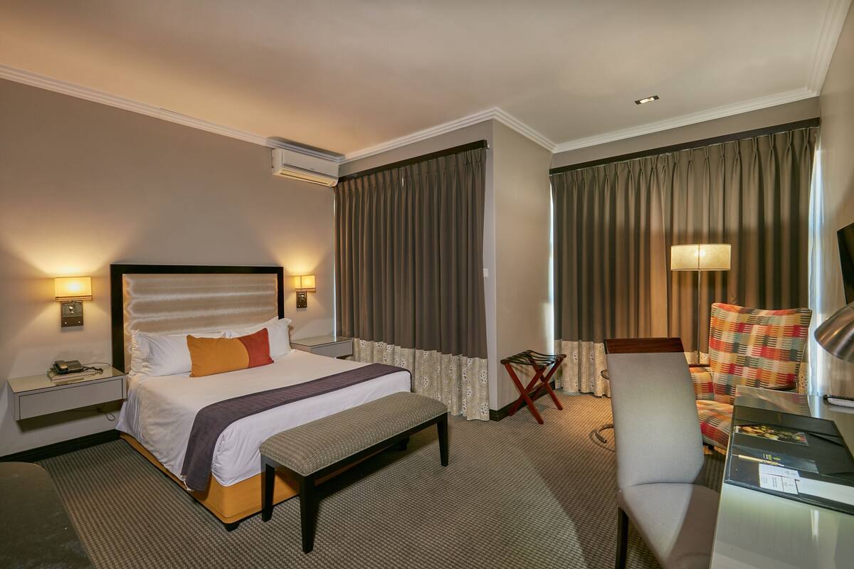 A hotel room with a bed, bedside table, lamp, chair, and a desk with a lamp.