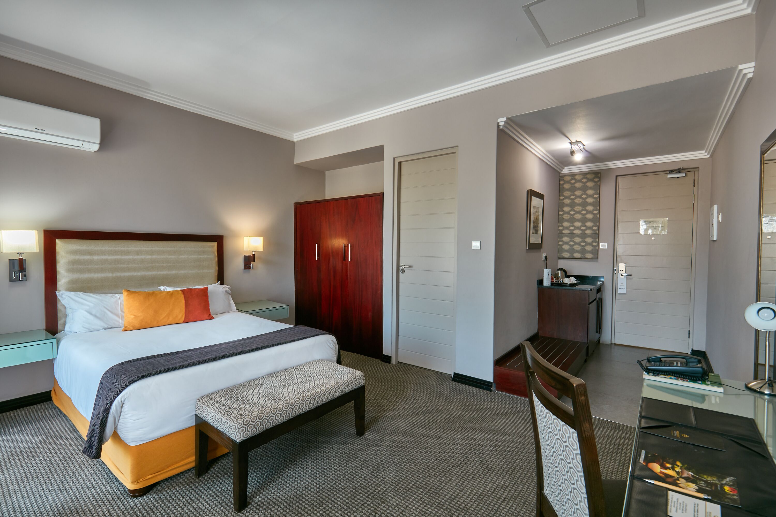 A hotel room with a large bed, bedside tables, a bench, a desk, and a wardrobe.