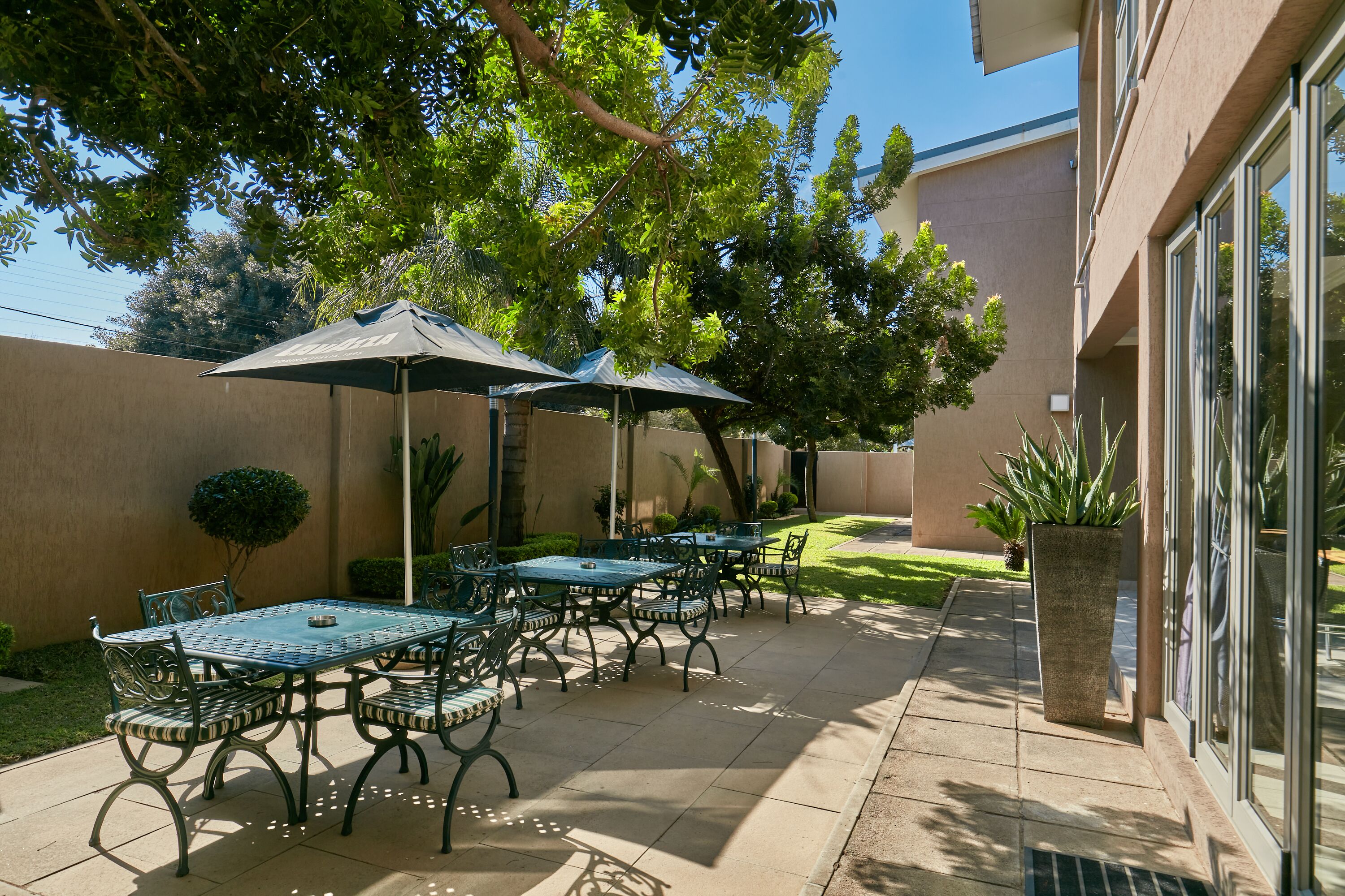 An outdoor patio with a table and chairs, surrounded by a low wall and a large tree.