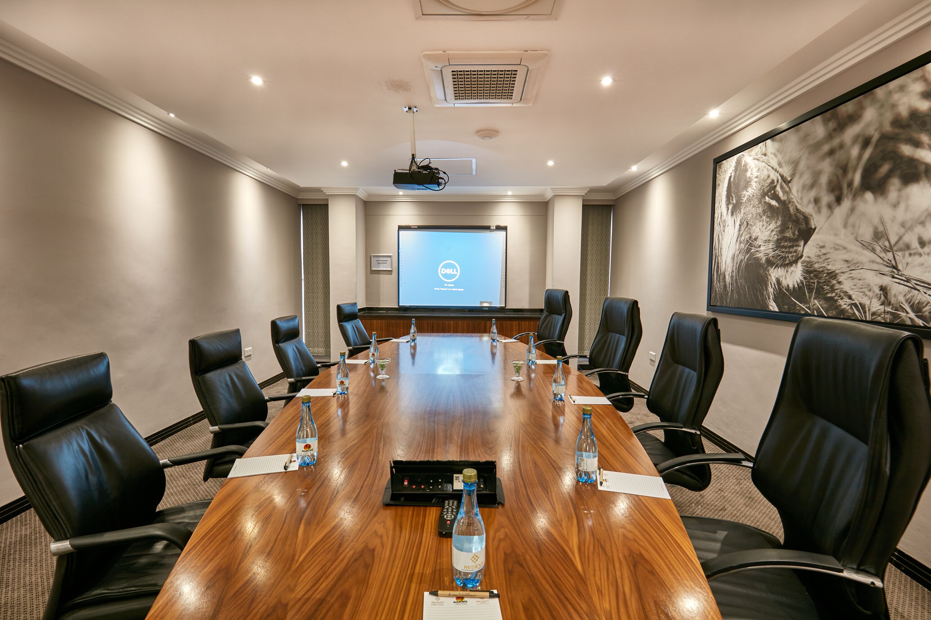 A conference room with a long wooden table, black leather chairs, a projector screen, and a large framed picture on the wall.