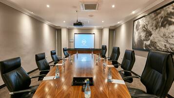 A conference room with a long wooden table, black leather chairs, a projector screen, and a large framed picture on the wall.