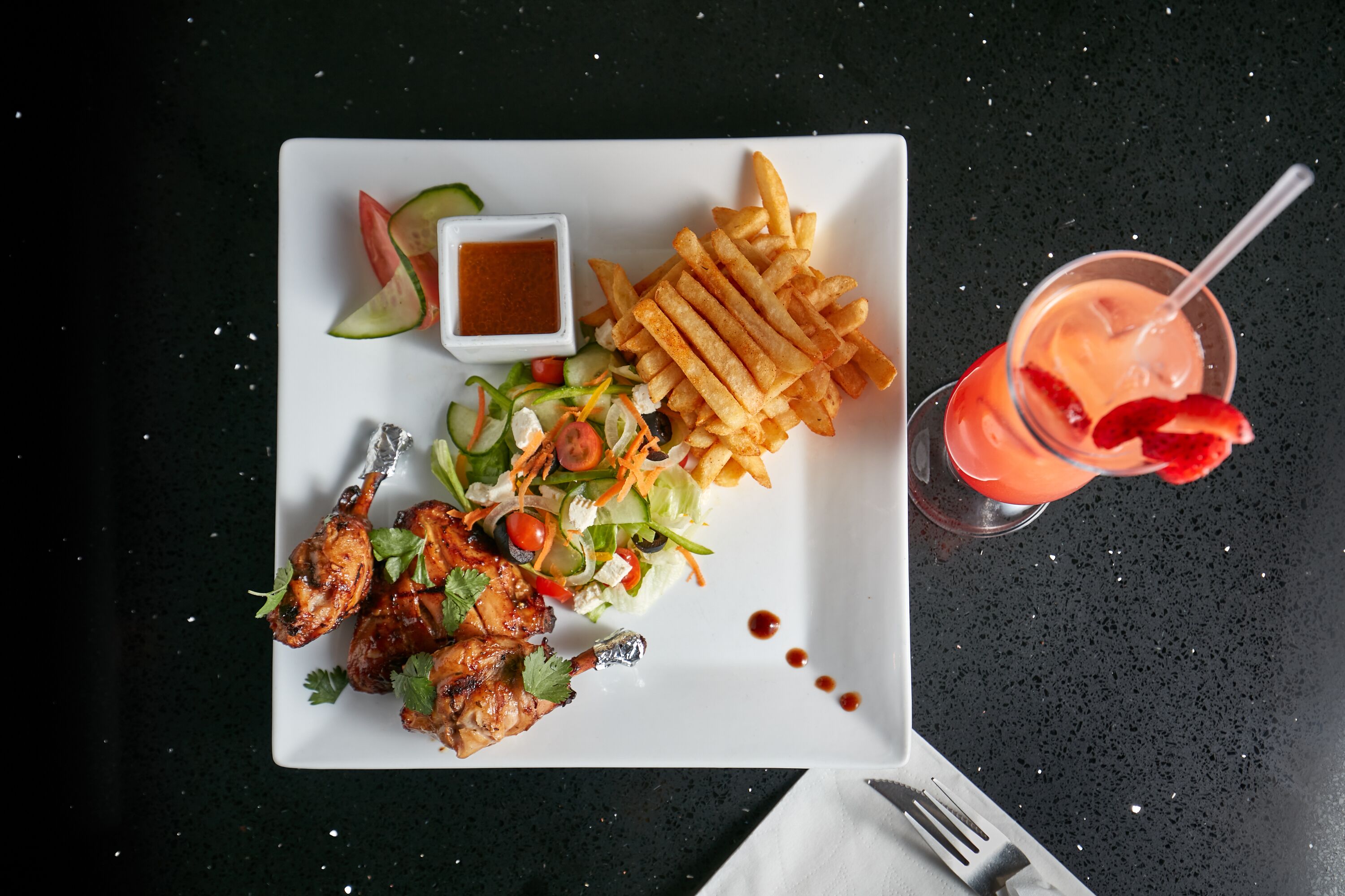 Ein Teller mit gegrilltem Hähnchen, Pommes, Salat und einem Cocktail.