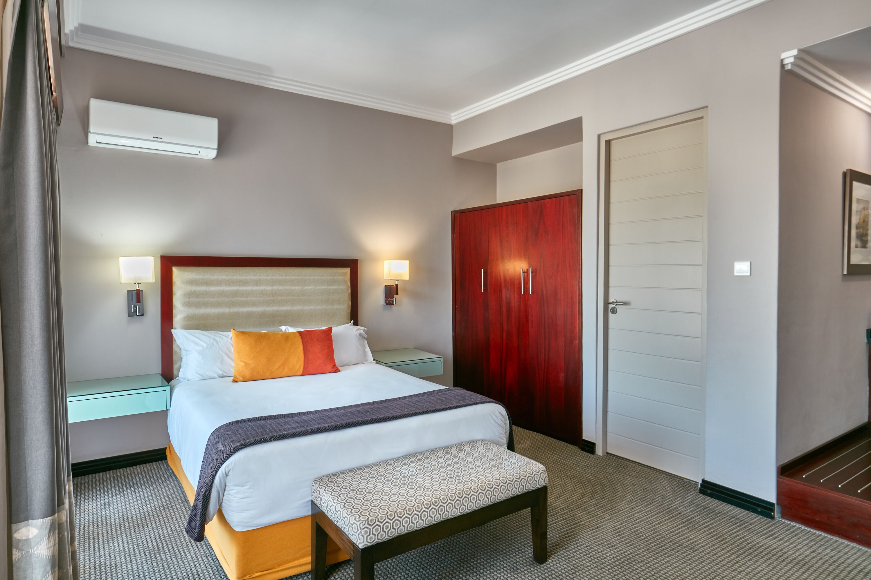 A hotel room with a bed, bedside tables, a wardrobe, and an air conditioning unit.