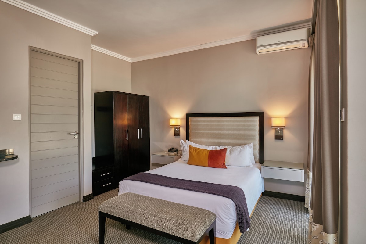 A hotel room with a bed, bedside tables, a wardrobe, and an air conditioning unit.
