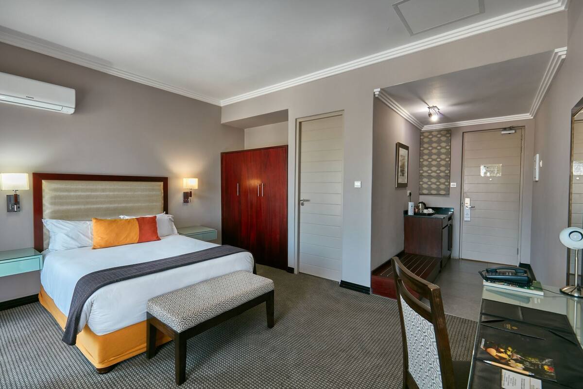 A hotel room with a bed, bedside table, lamp, wardrobe, desk, chair, and a mirror.