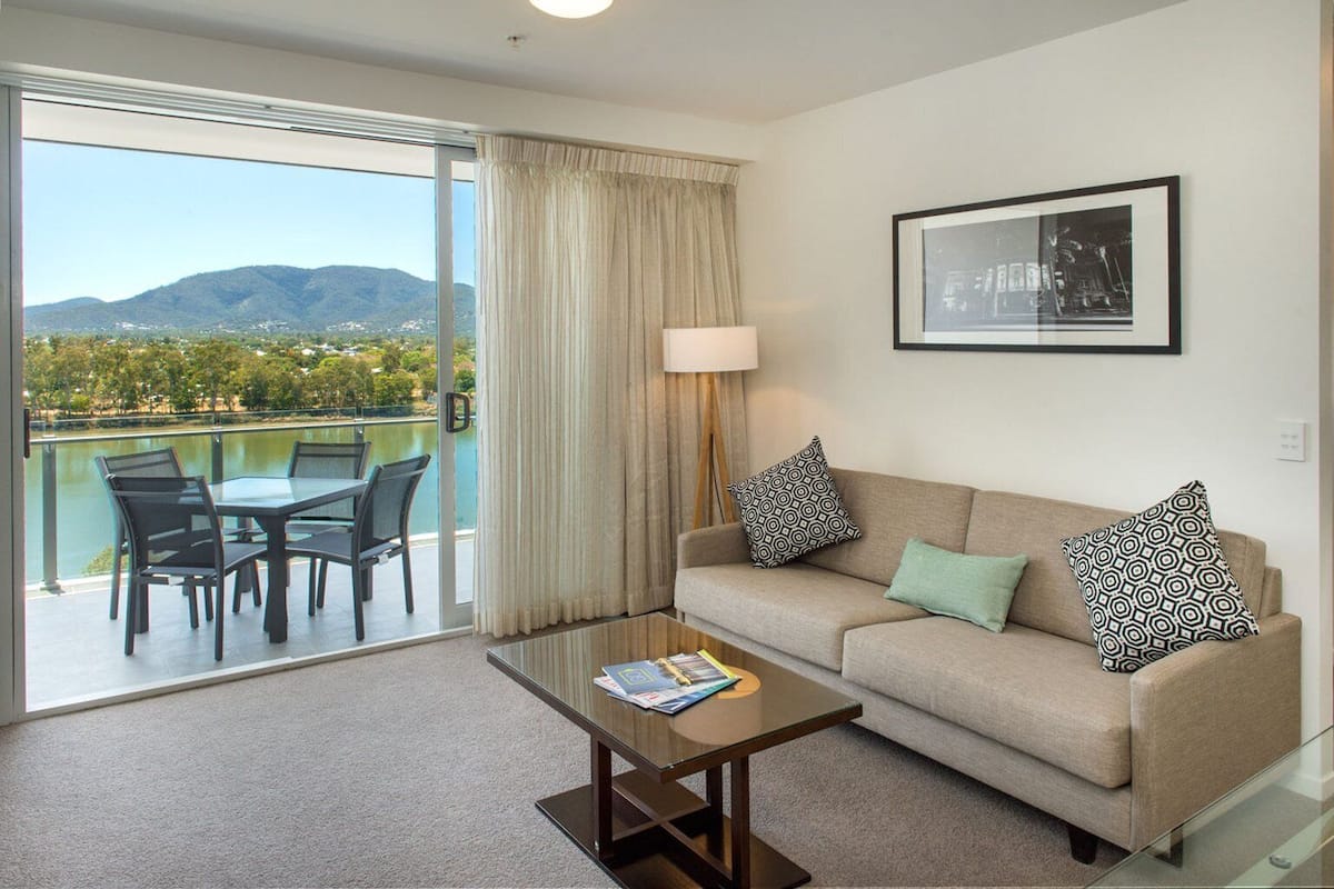 Ein Hotelzimmer mit Balkon, einer Couch mit gemusterten Kissen, einem Glastisch und einem gerahmten Bild an der Wand.