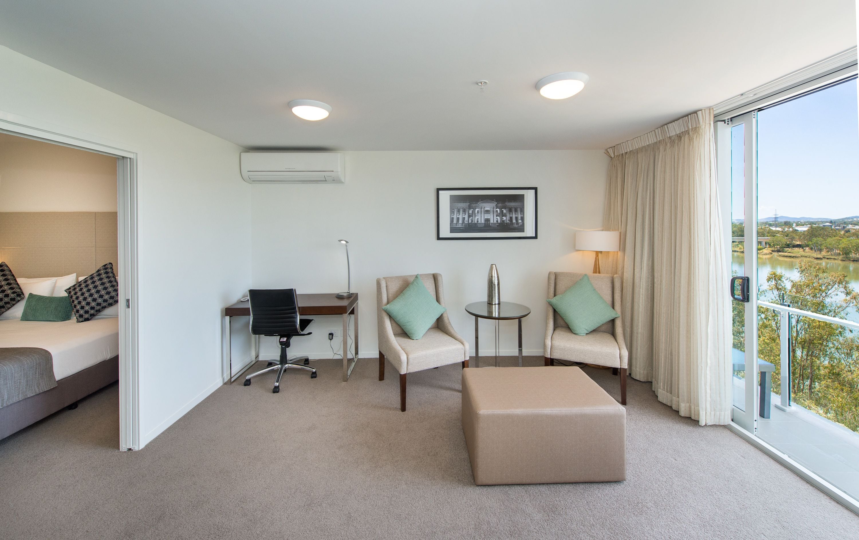 Una habitación de hotel con cama, dos sillones, un escritorio, una silla, una mesita, una lámpara y vistas a una masa de agua.