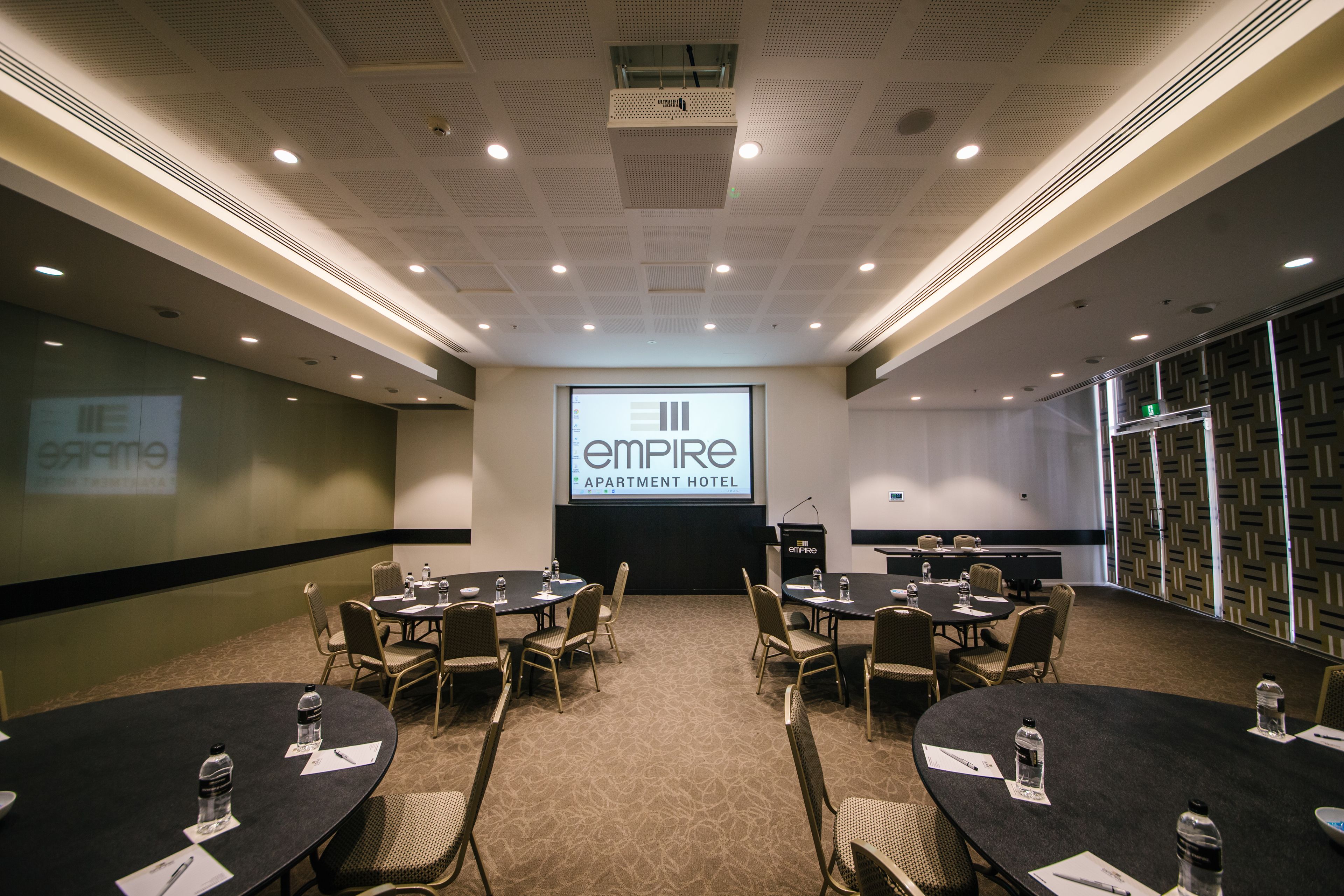 Un salón de conferencias con varias mesas y sillas, una pantalla de proyector mostrando 'Empire Apartment Hotel' y un atril con un micrófono.