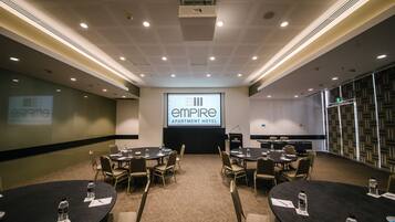 Un salón de conferencias con varias mesas y sillas, una pantalla de proyector mostrando 'Empire Apartment Hotel' y un atril con un micrófono.