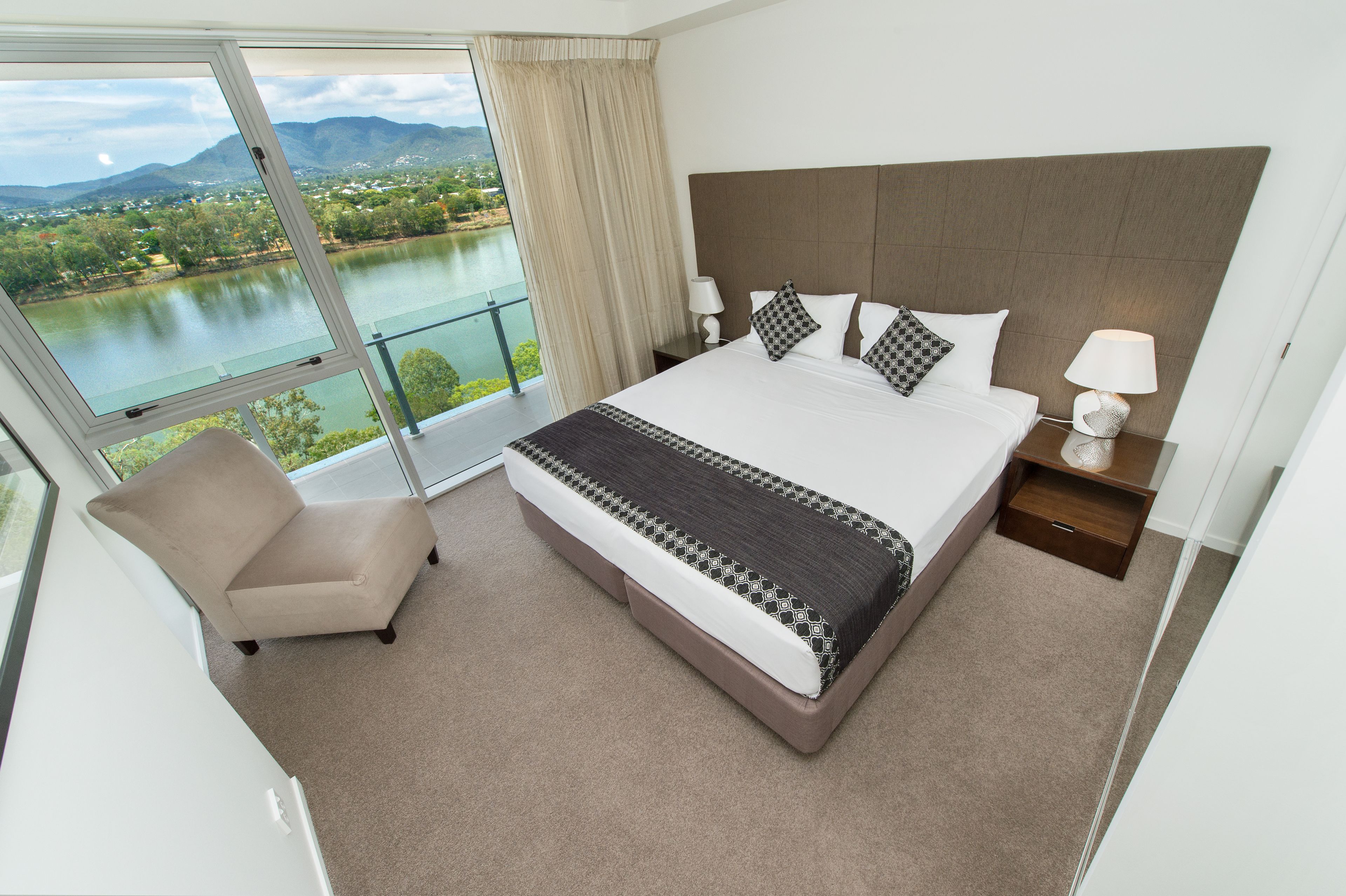 A large bed with a headboard, a bedside table with a lamp, a chair, and a view of a lake and mountains through a window.