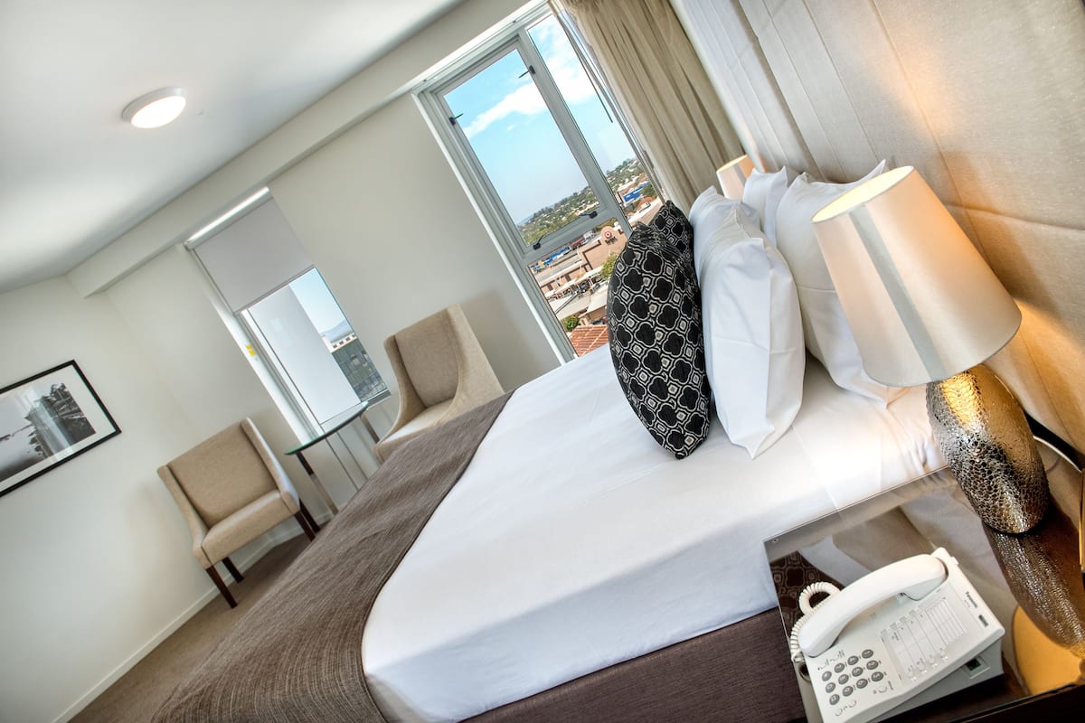 Ein Hotelzimmer mit einem Bett, einem Telefon, einem Sessel, einem Fenster mit Blick auf die Stadt und einem gerahmten Bild an der Wand.