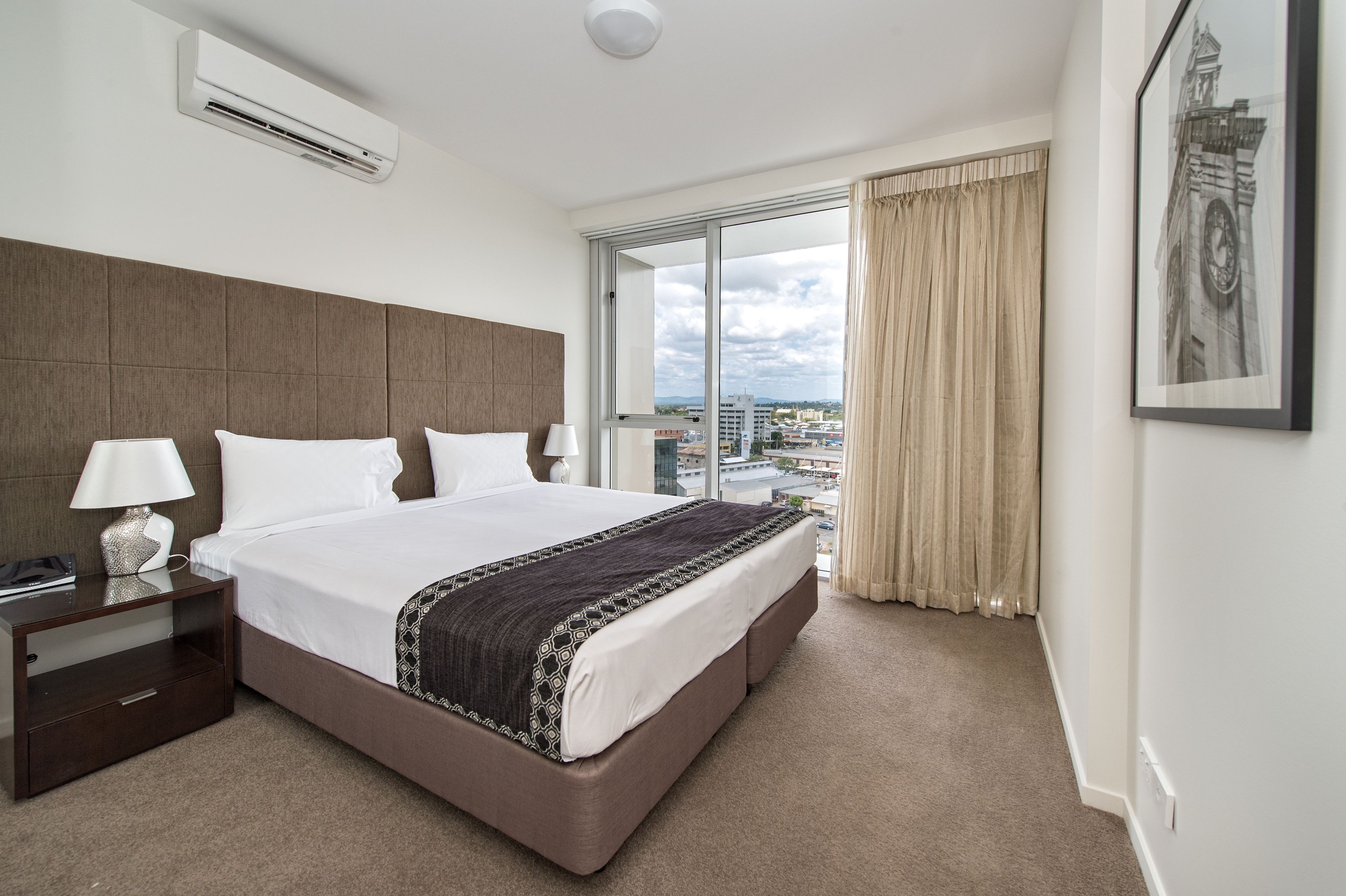A hotel room with a large bed, a nightstand, a lamp, an air conditioner, a large window with curtains, and a framed picture on the wall.
