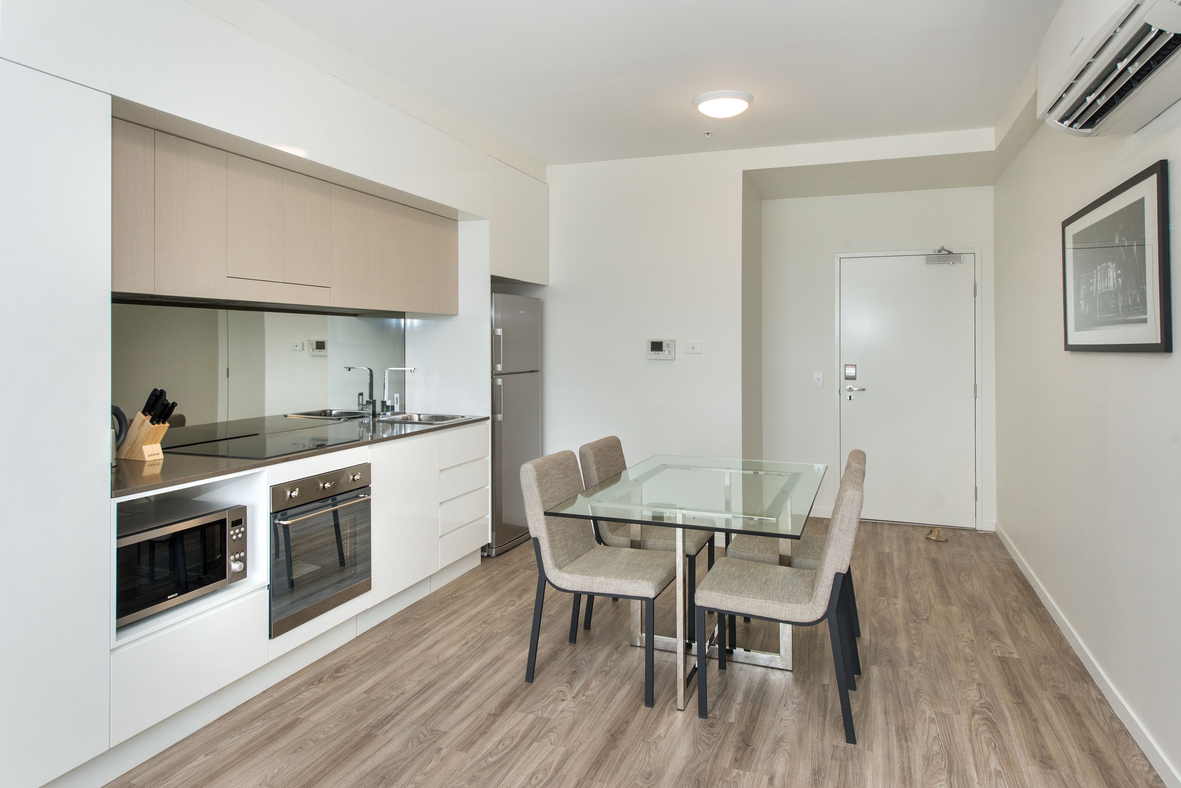 A modern kitchen with built-in appliances, a dining area with a glass table and chairs, and a door leading to another room.