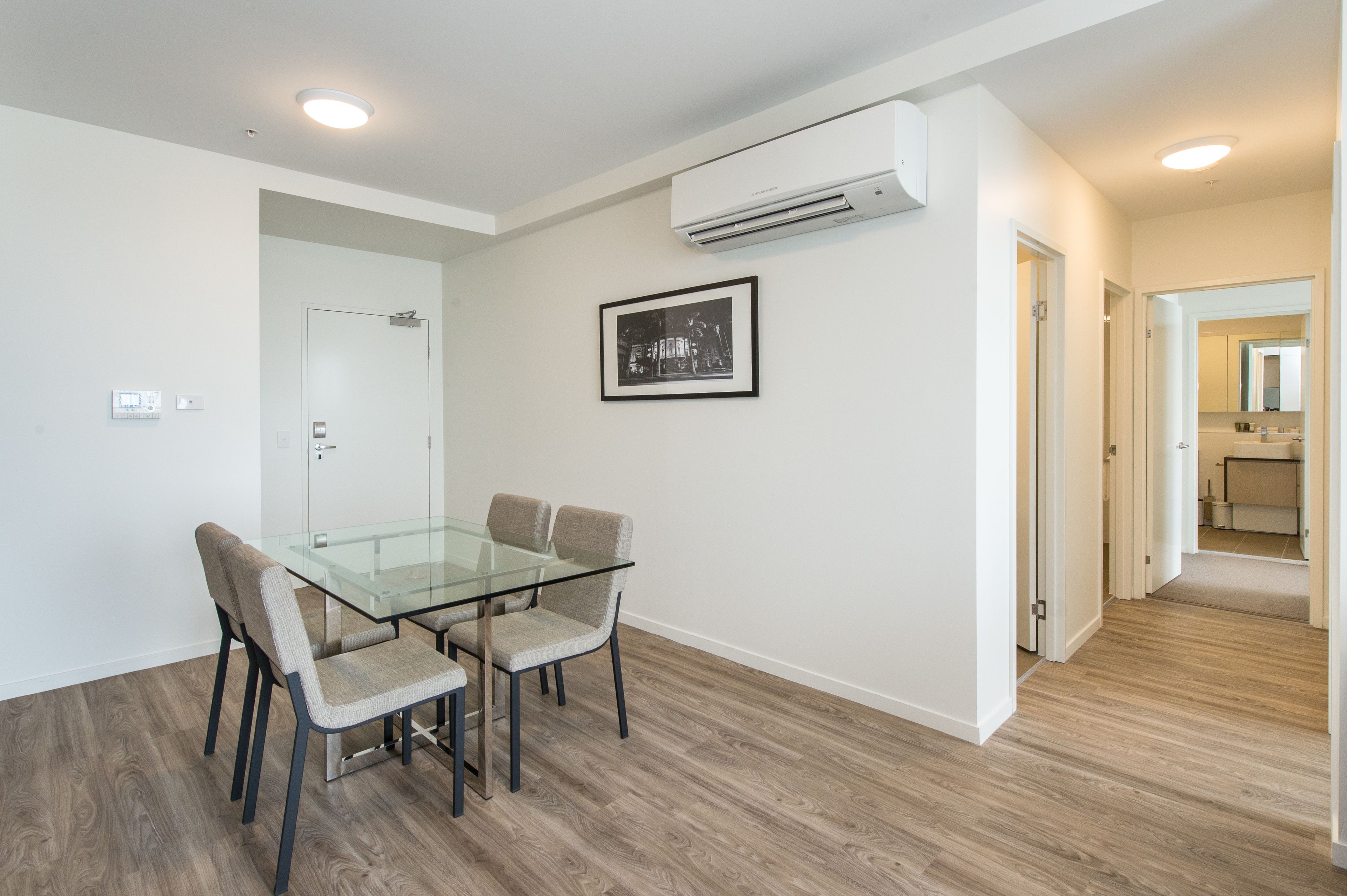 A modern room with a glass dining table, four chairs, a white door, an air conditioning unit, and a framed picture on the wall.