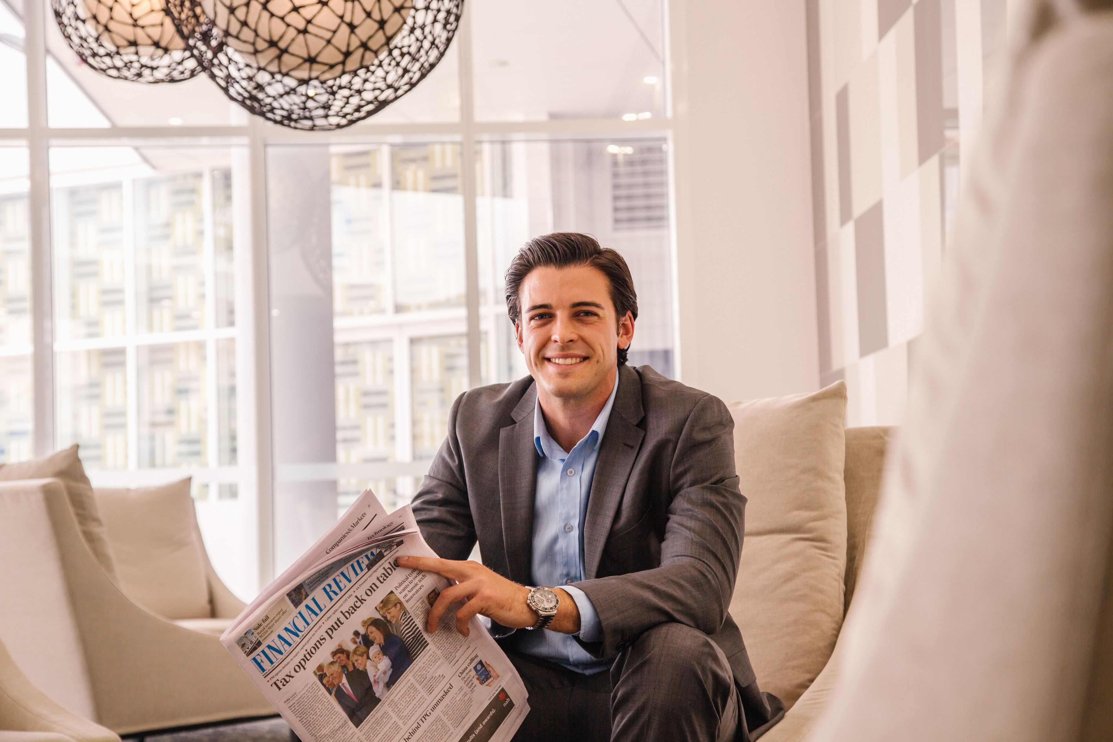 A man sitting on a sofa reading a newspaper in a modern room with large windows and a geometric light fixture.