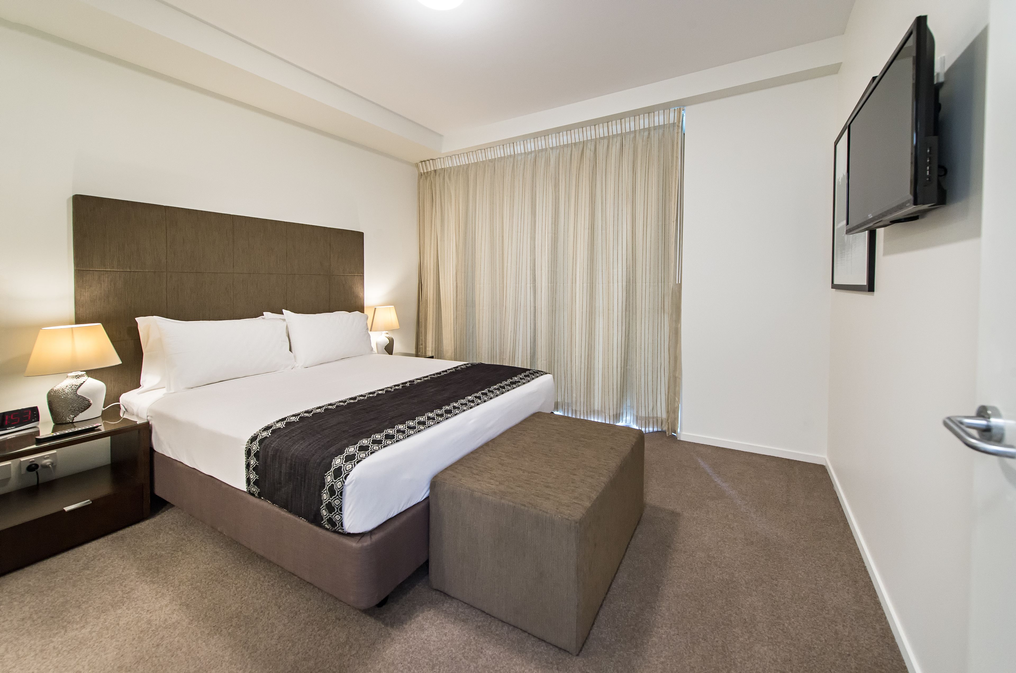 A hotel room with a bed, bedside table, lamp, TV, and curtains.