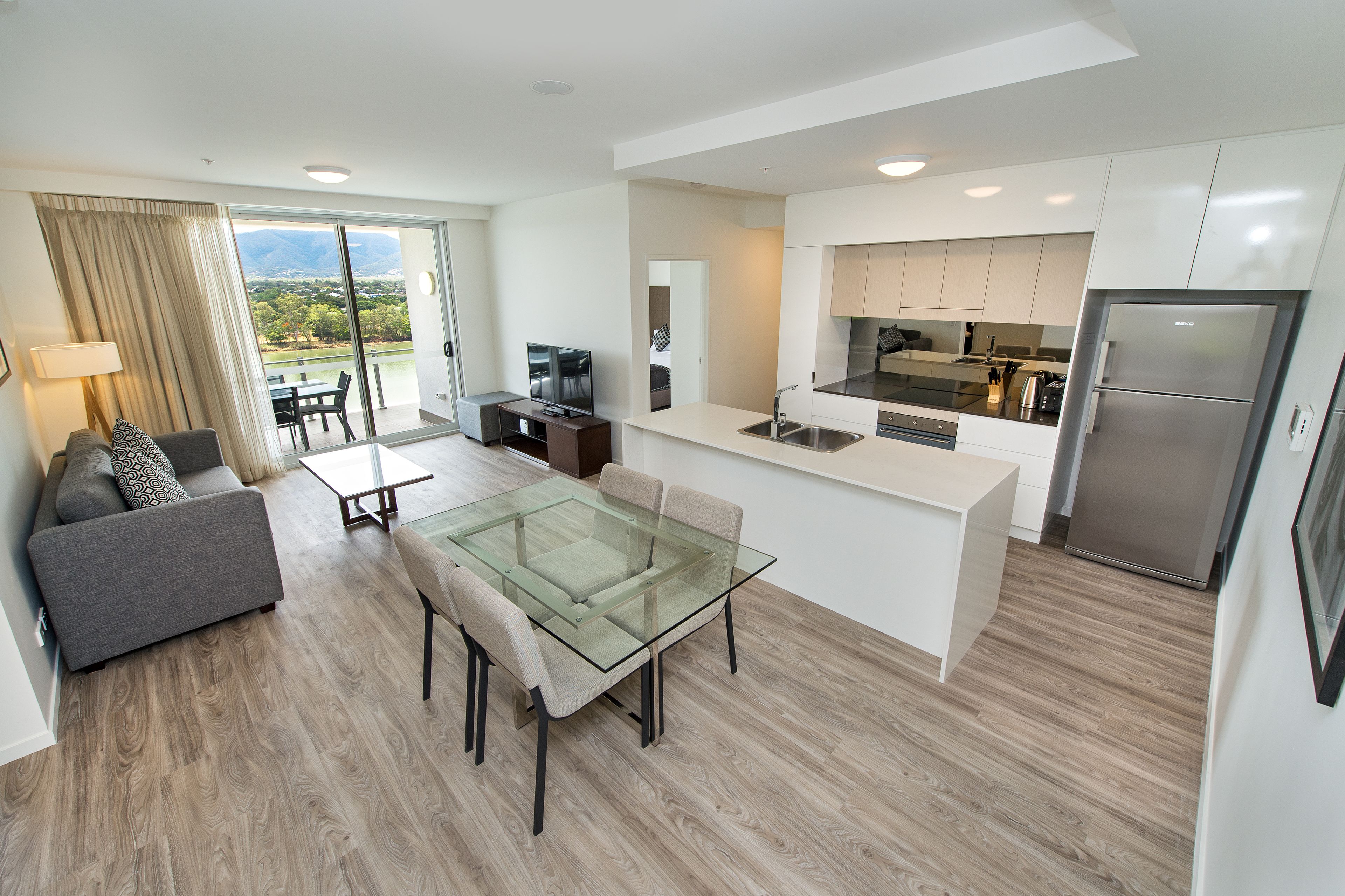 A modern apartment with a glass dining table, grey sofa, and a kitchen with a stainless steel refrigerator.