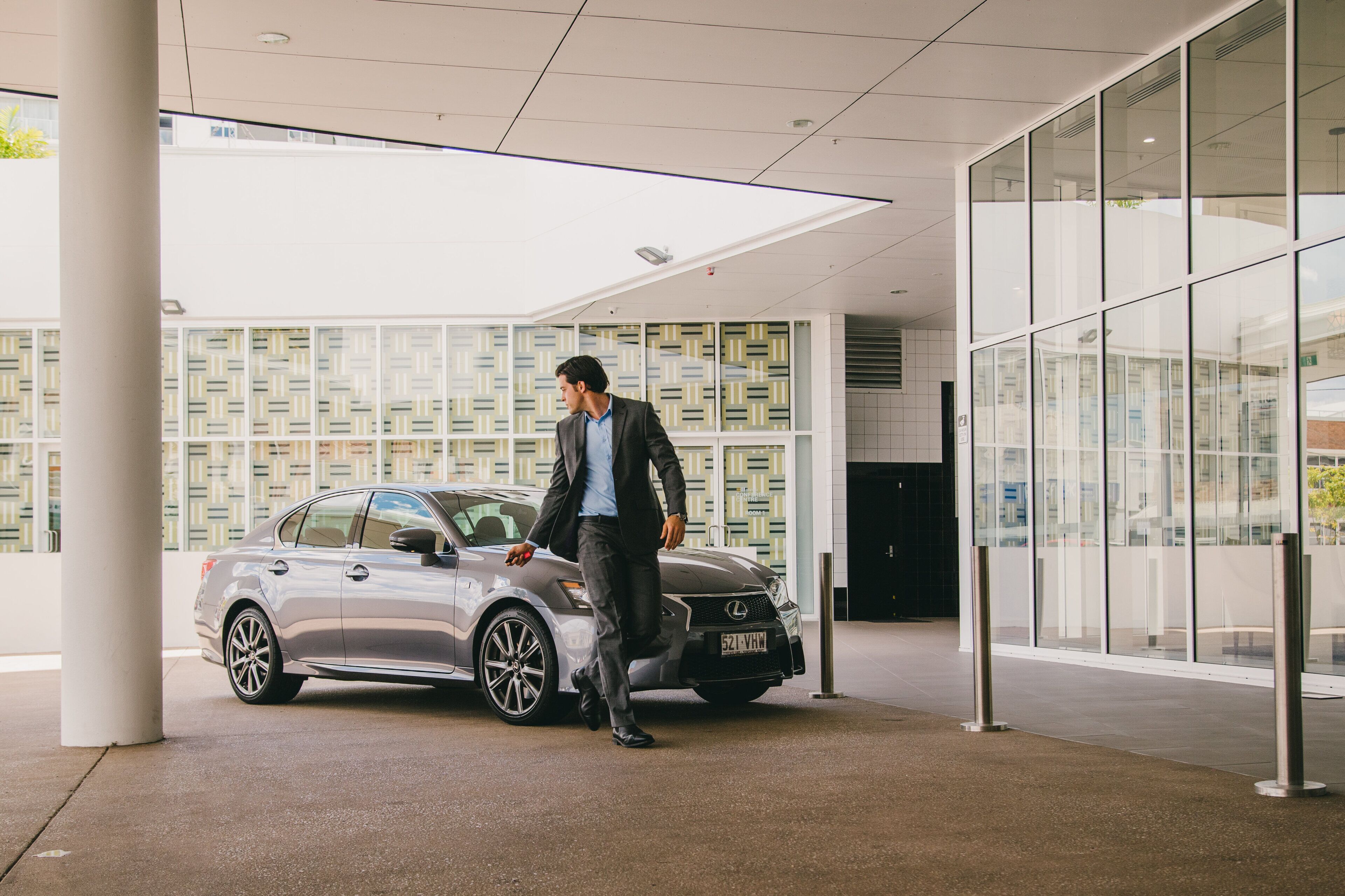 Un homme en costume marchant à côté d’une voiture Lexus argentée, devant un bâtiment moderne doté de grandes fenêtres et de murs à motifs.