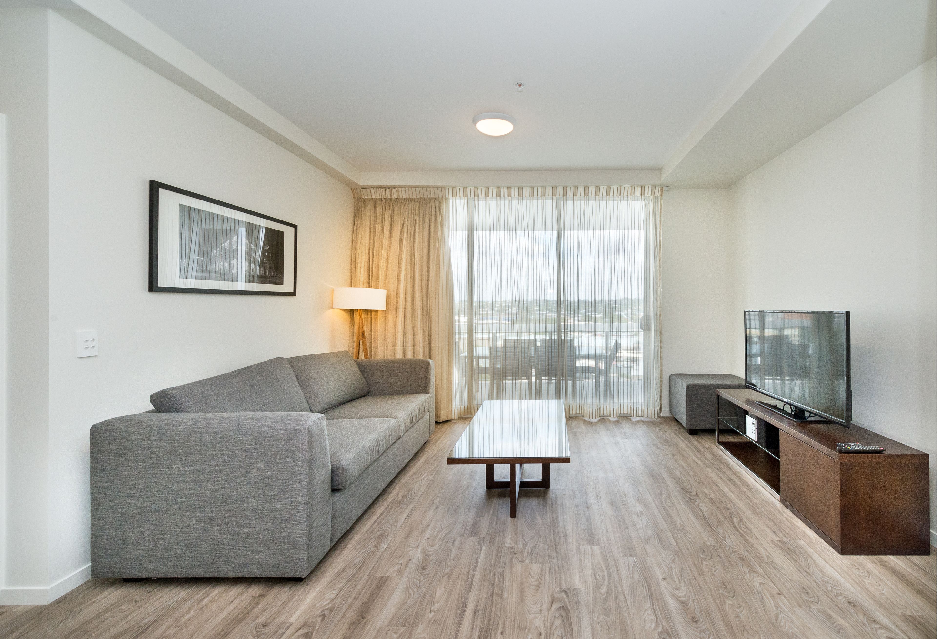 Un salón moderno con un sofá gris, una mesa de centro de madera, un televisor sobre un mueble de madera y un amplio ventanal con cortinas de encaje.