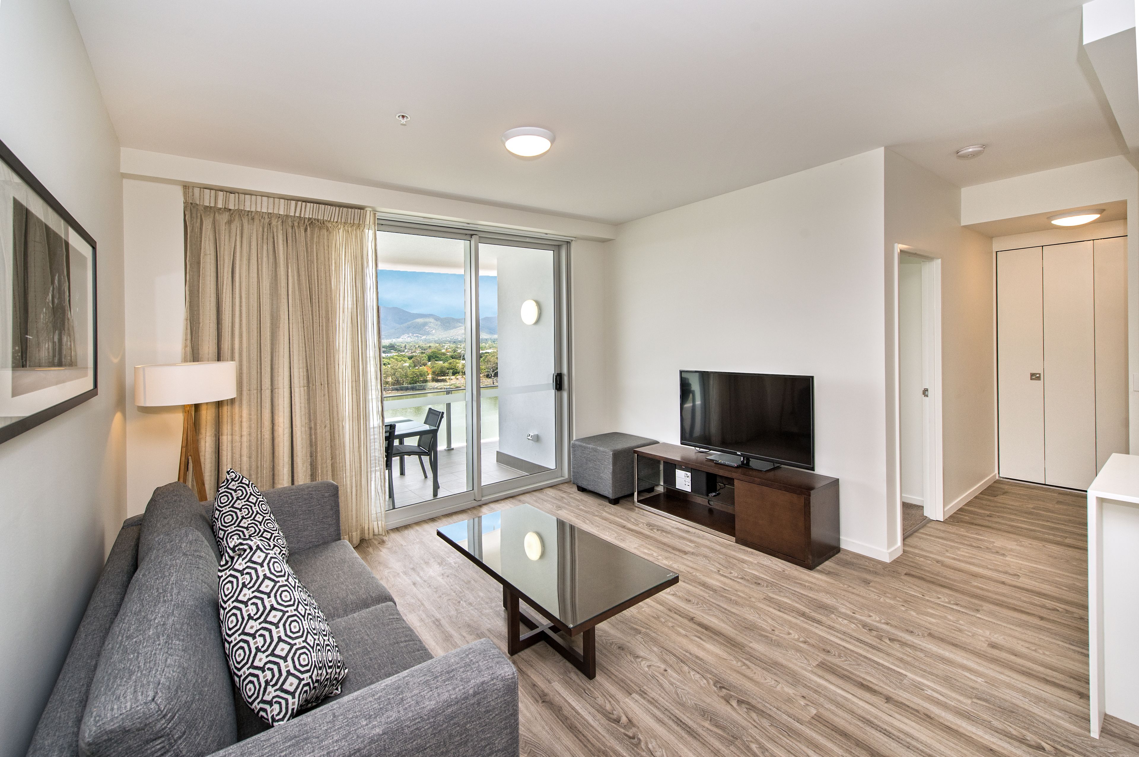 A modern living room with a sofa, coffee table, and TV. It features a sliding glass door leading to a balcony with a city view.