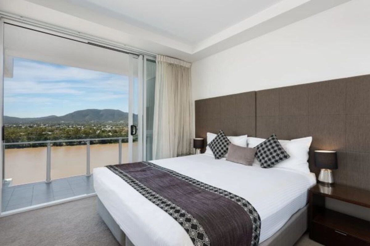 A hotel room with a large bed, a nightstand, a window with a view of a body of water and mountains, and a balcony.