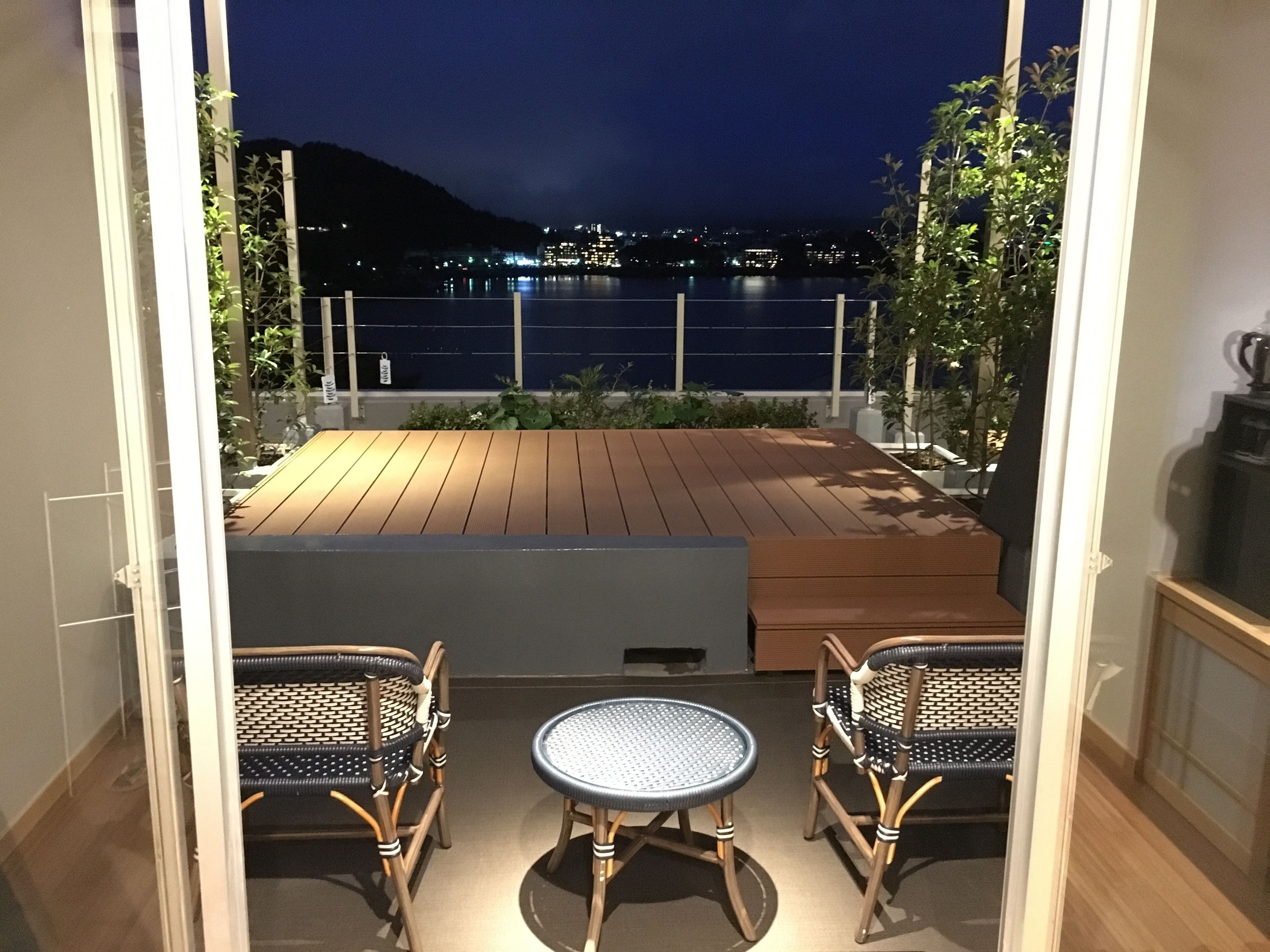A balcony with wooden flooring, two wicker chairs, a small round table, and a view of a body of water at night.