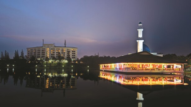 A waterfront hotel with a reflection in the water, a prominent building with a tower, and a boat docked nearby. - RHR Hotel at Uniten (Kajang)