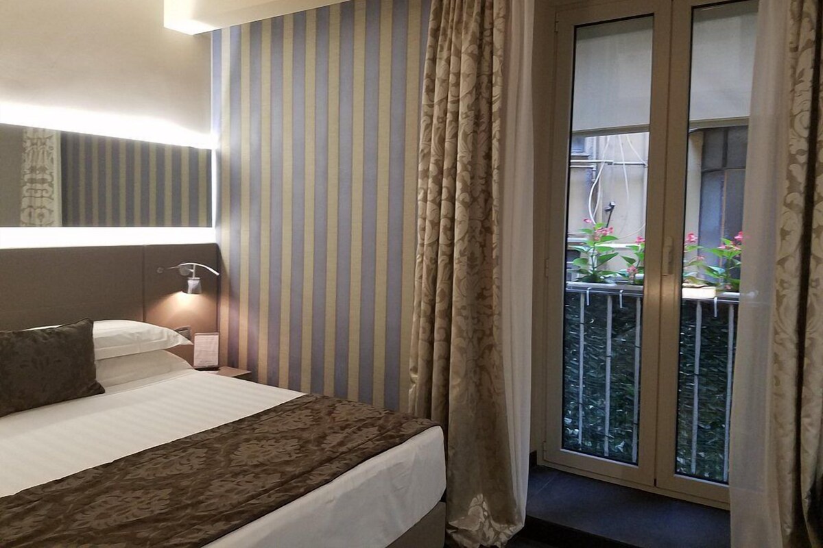 A hotel room with a bed, a bedside table, a lamp, striped curtains, and a view of a balcony with plants.