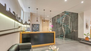 A modern lobby with a reception desk, a black leather sofa, a glass coffee table with a vase, a staircase with a metal railing, and a cityscape mural on the wall. - Trevi Palace Hotel (Rome)