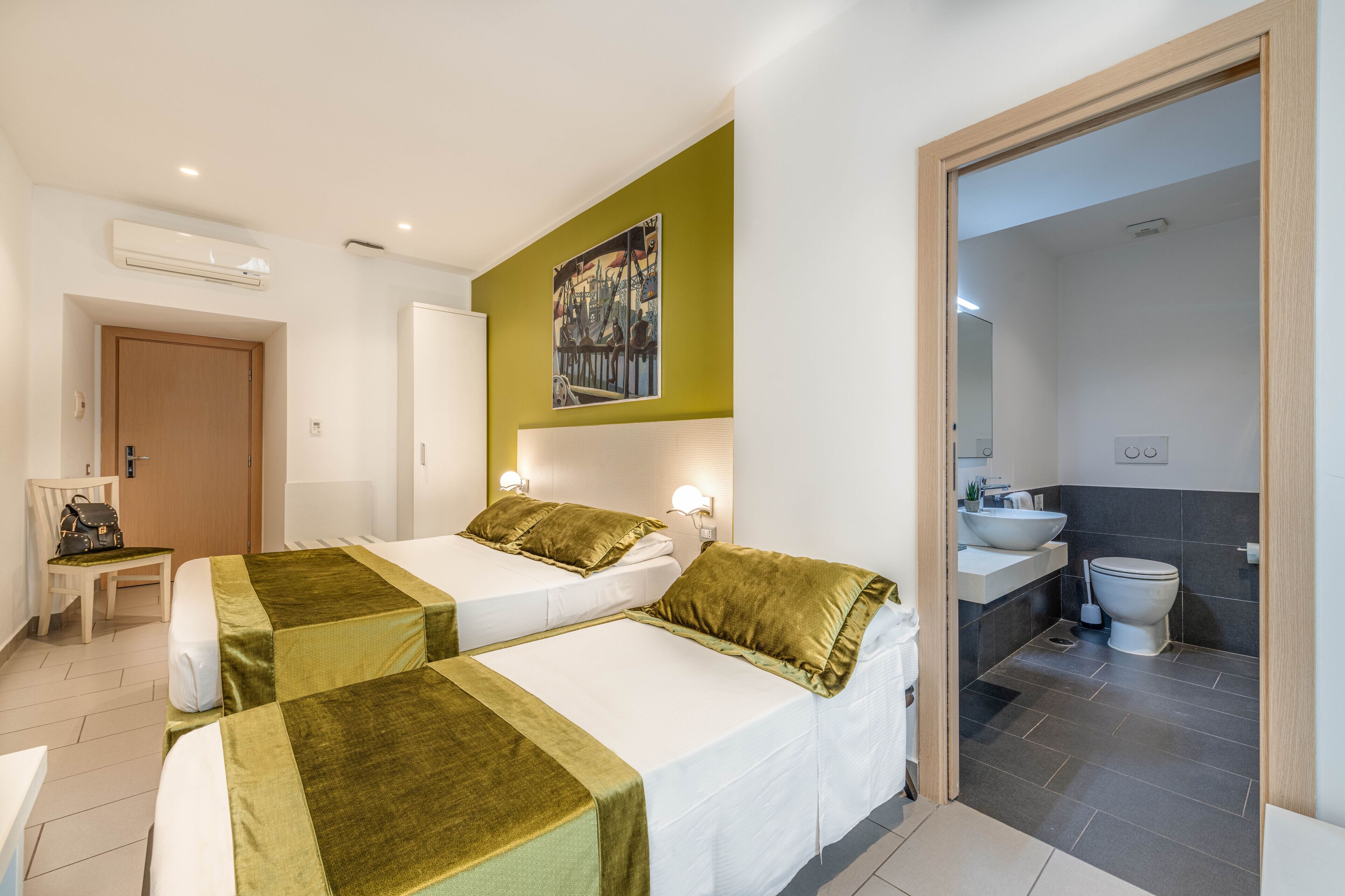 Una camera d'albergo con due letti, un quadro appeso al muro, un bagno con lavandino e WC, e una sedia con una borsa.