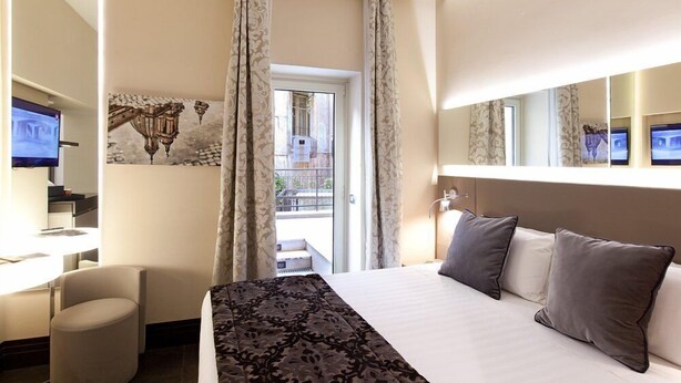 A hotel room with a bed, a desk with a computer, a chair, a large window with curtains, and a balcony view. - Trevi Palace Hotel (Rome)