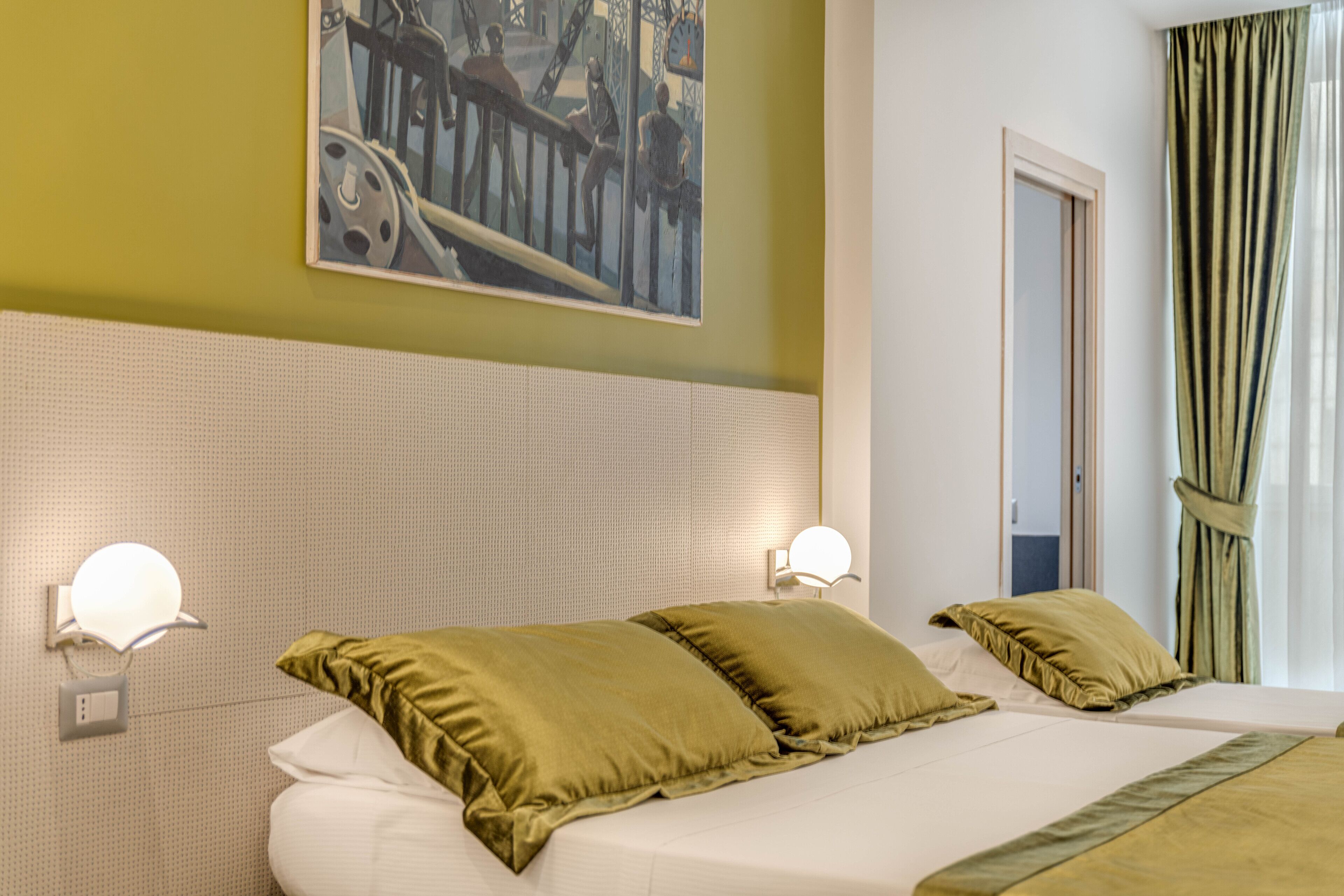 Una camera d'albergo con un letto grande, due abat-jour, un quadro appeso al muro e una finestra con tende.