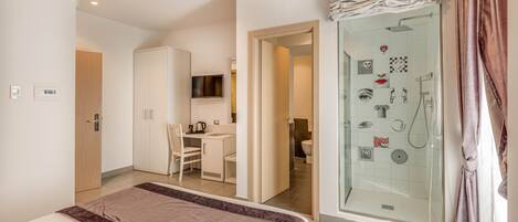 A hotel room with a bed, a desk, a wardrobe, a shower with a glass door, and a television mounted on the wall.