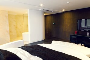 A modern hotel room with a large bed, a separate sitting area, a shower, and a dark wooden wall.