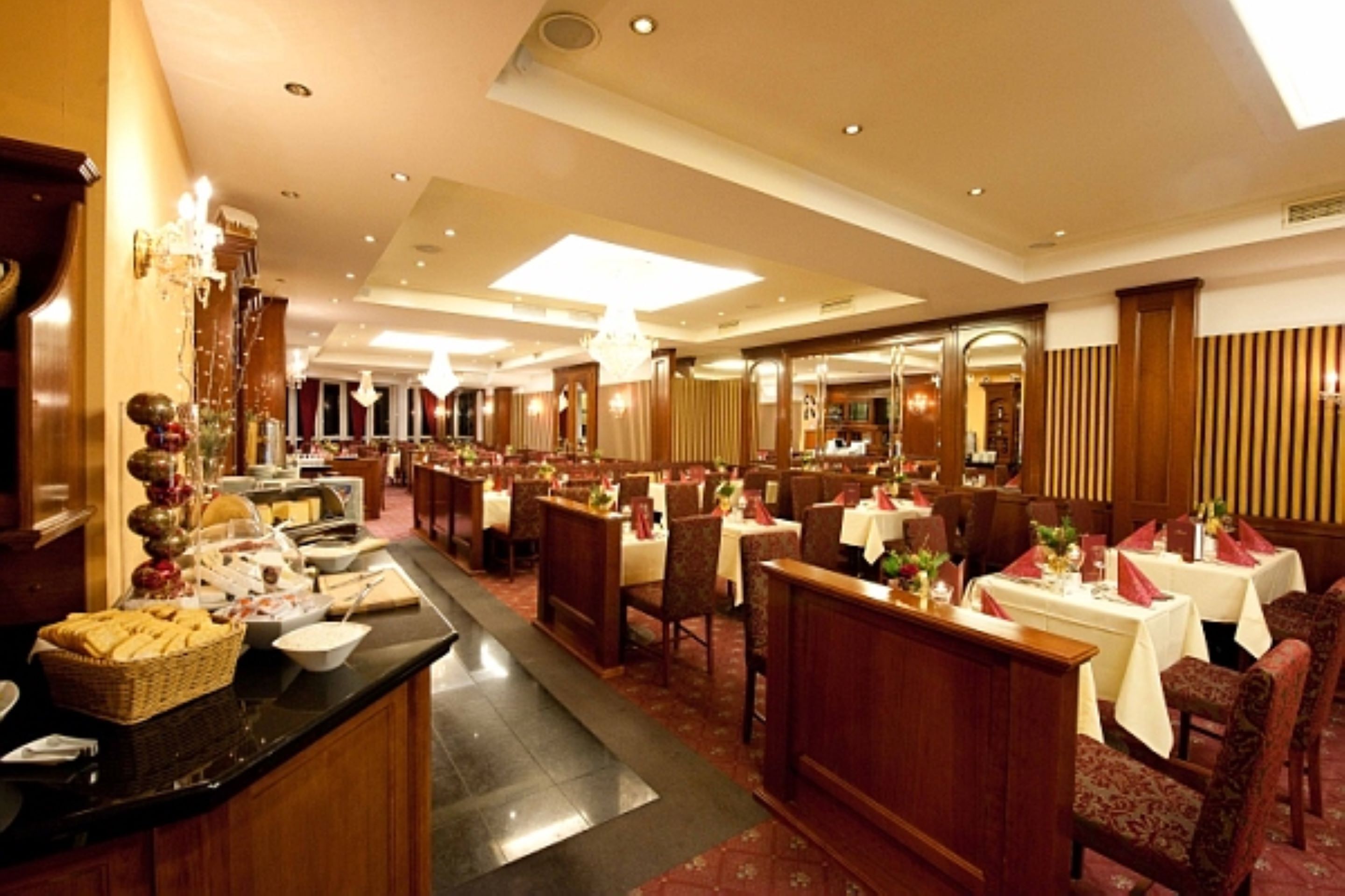 A spacious hotel dining area with multiple tables and chairs, a buffet counter, and a large chandelier.