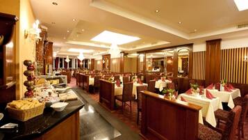 A spacious hotel dining area with multiple tables and chairs, a buffet counter, and a large chandelier.